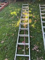 Photo showing two aluminum extension ladders laid on grass outdoors, tied with red ropes to secure sections.