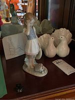 Figurines displayed on wooden surface with mirrored background, showing 'Dropping The Flowers' girl figurine and Couple of Doves lovebirds, alongside Lladro plaque.