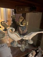 Three Lladro porcelain figurines displayed on a glass shelf under warm lighting: a girl holding a flower pot, a white duck, and a preening goose.