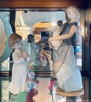 Two porcelain figurines displayed on shelf with mirror background showing back details. One figurine is a girl holding a bunny, the other a praying child with toy teddy bear.