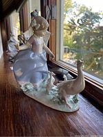 Porcelain figurine showing the left side profile of a young girl with geese and goslings, glossy finish, positioned near a window with natural light highlighting details.