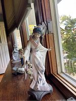 Front angled view of the Lladro Japanese Geisha porcelain figurine placed on wooden ledge by the window, showing details of kimono, posture, and facial expression.