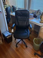 Front view of black faux leather office chair positioned on wooden floor near desk and window, showing padded armrests and cushioned seat and backrest.