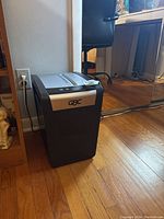 Photo of the black and silver GBC ShredMaster PSX10-06 paper shredder standing on a wooden floor next to a wall and desk.