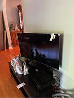 TV with screen turned off, placed on dark wooden media table with some figurines nearby. Tags on TV visible indicating auction item.