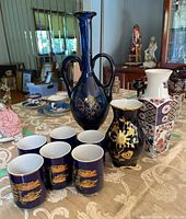 Group photo showing the tall cobalt blue vase with two handles, smaller cobalt blue vase, white patterned vase, and six matching cobalt blue cups with gold decorations arranged on a table.