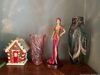 Four items on shelf: ceramic gingerbread house, smaller red-pink art glass vase, female figurine, and large multicolor art glass vase
