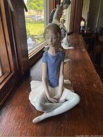 Front view of the Lladro Beth sitting ballerina figurine in light blue top and white skirt on wooden surface by window.