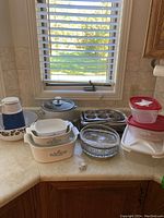 Wide shot depicting the majority of the lot items including CorningWare dishes, plastic storage containers, muffin pan, clear glass dish, and electric rice cooker