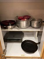 Wide shot of shelf containing pots with lids, frying pan with metallic handle, two broiler trays, cast iron pan, and oven mitts on top of pots.