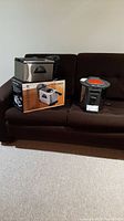 Two deep fryers on a dark brown couch: one stainless steel HomeMax fryer with box, and a gray SuperFryer with orange top.