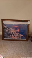 Framed poster of the Grand Canyon leaning against a wall on carpet. The image shows the canyon with rock formations and colorful lighting.