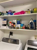 Wide shot of two white shelves above a washer and dryer, containing multiple boxes of light bulbs from Philips and other brands, tape measure, iron, and other small packaged items.