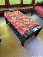 Dark brown woven wicker rectangular ottoman with red cushion featuring beige leaf patterns placed indoors on tiled floor.