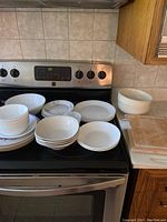 Photo of white plates and bowls, stacked and arranged on stove and countertop.