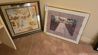 View of two framed prints placed on the carpeted floor, showing the floral still life print with mirrored border and the garden path print with pink floral matte, both with gold frames.