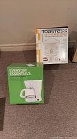 Both unopened original boxed items: Toastess Jar Style Rice Cooker and Everyday Essentials 12 Cup Coffee Maker seen side by side on floor.