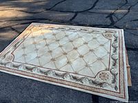 Full view of the beige wool area rug positioned outdoors on pavement, showing decorative border and central floral pattern, partially folded to reveal non-slip underlay underneath.