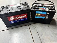 Two large marine deep cycle batteries side by side on floor: Duralast on left, EverStart on right.