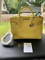 Front view of yellow Michael Kors purse, showing handles, MK logo charm, and separate shoulder strap wrapped in protective paper.