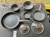 Full cooking set displayed on black surface outdoors: 3 frying pans, 1 saucepan, 1 cast iron skillet, 2 glass lids.