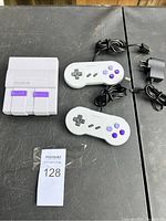 Photo showing the compact retro gaming console, two wired controllers with purple buttons, and the power adapter.