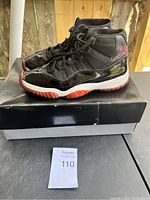Pair of black and red Jordan 11 Retro Bred shoes placed on a box, showing side view and detailing including Jumpman logo on heel.