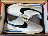 Side view of new condition white Nike Cortez shoes with black swoosh and blue midsole stripe inside original box.