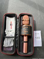 Wireless karaoke microphone in rose gold metallic finish with mesh grille speaker section and control buttons, placed inside a black zippered hard carrying case. Case includes mesh pocket containing user manual and charging cable.