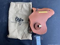 Brown leather purse with unique shape, zipper pocket, brass Orijin Culture nameplate, rolled strap, and branded fabric storage bag shown on black surface.