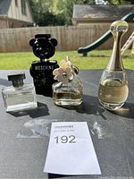 Main photo showing four perfume bottles on outdoor table: Moschino black bear, Marc Jacobs Daisy, Dior J'adore, and Jean Paul Gaultier