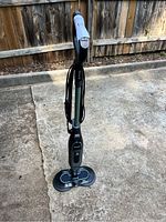 Full view of a Shark upright steam mop in black and teal, standing on concrete flooring, showing ergonomic handle and power cord wrapped around body.