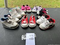 Lot of eight toddler shoes in various styles and sizes arranged on a black surface outdoors. Includes white and blue Air Jordan sneakers, red and white canvas sneakers, white sneakers with red details, and pink and purple slip-on shoes.