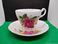 White ceramic tea cup with floral design sitting on matching saucer, displayed against green background.