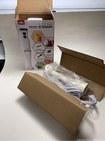 Electric hand blender in box with blending shaft and cord.