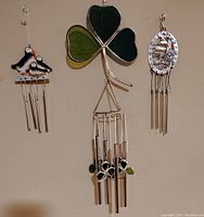 Three wind chimes hanging vertically on a wall. Left small chime features puffins and four metal tubes. Center large chime has a three-leaf stained glass shamrock top with five tubular metal chimes and metal shamrock charms. Right small chime shows an oval metallic top with five tubular metal chimes below.