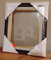 Front view of the picture frame wrapped in plastic with protective white cardboard corners showing dark wood and gold trim.