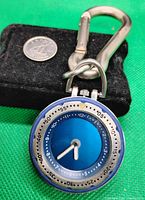 Front view of the pocket watch displaying its vibrant blue dial with hour and minute hands, attached to a silver carabiner clip. A small black pouch and a quarter coin shown for scale.