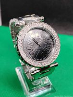 Front view of silver-tone wristwatch with a black dial and rhinestone-encrusted bezel showing the time near 12 o'clock position.