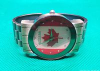 Front view of metal quartz watch with Canadian flag design on the face, visible 'ecosse' text, and metal linked band on green background.