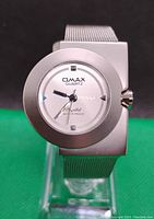 Front view of Omax Quartz watch showing round dial within a square bezel and silver mesh strap.