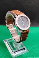 Side angled view of quartz watch showing round silver case, grey face with Roman numerals, brown leather strap with white stitching, and buckle closure