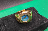 Close-up of a gold-tone finger watch with a blue dial placed on black fabric.