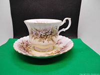 Front view of vintage Royal Albert tea cup with floral design on cream background and gold trim on handle and rim, with matching saucer beneath showing scalloped edge.