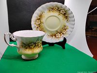 Photo showing the tea cup in front of the matching saucer standing on a black stand against a green and white background; floral pattern and gold accents visible.