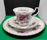 Front view of stacked tea cup, saucer, and small plate showing floral design and gold trim. Tea cup has March inscribed inside.
