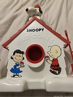 Front view of Snoopy Sno-Cones maker showing house shape with red roof, Snoopy figure on top with red hat, and Charlie Brown and Lucy graphics on front holding sour cone cups.
