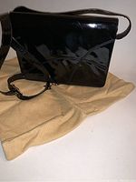 Black shiny patent leather purse positioned on light beige cloth, showing full front and strap.
