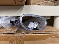 Single pair of clear plastic safety goggles on cardboard background, strap and wrap-around lens design visible.