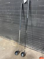 Two TaylorMade golf driver clubs leaning against a black wall, showing the club heads and shafts.